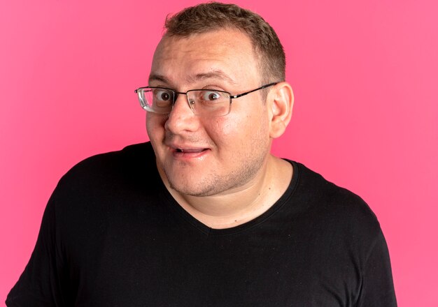 Homme en surpoids à lunettes portant un t-shirt noir avec un visage heureux souriant sur rose