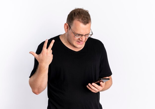 Homme en surpoids portant un t-shirt noir dans des verres en regardant l'écran de son smartphone confus et mécontent de faire des gestes avec la main debout sur un mur blanc