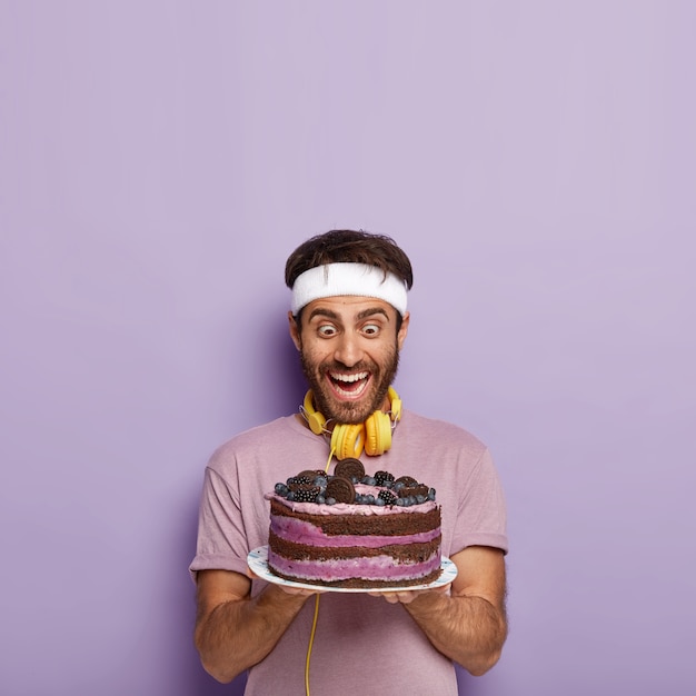 Un homme surpris et ravi regarde un délicieux gâteau, ressent la tentation, a faim après une séance d'entraînement active, porte un t-shirt décontracté, porte des écouteurs