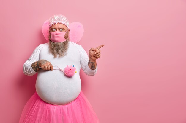 Un homme surpris se trompe tout en restant à la maison pendant la quarantaine pendant une longue période, porte un masque de protection, un costume de fée, indique sur l'espace de copie, montre une chose étonnante