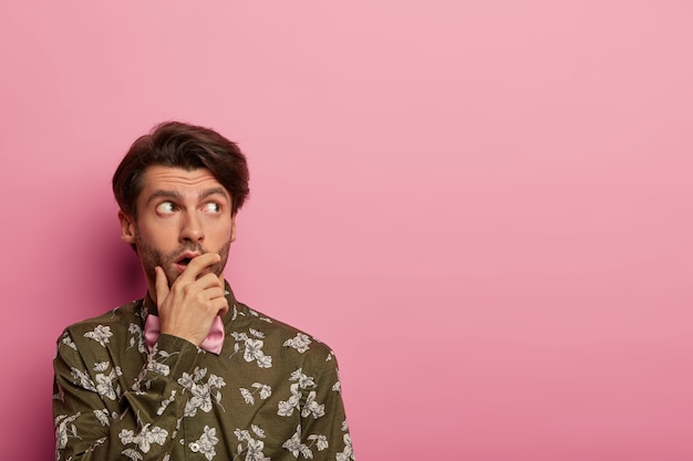 Photo gratuite un homme surpris et songeur couvre la bouche, concentré sur le côté, réfléchit à la révélation choquante, vêtu d'une chemise à fleurs