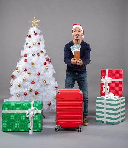 Homme surpris avec valise rouge tenant ses billets de voyage avec les deux mains près de l'arbre de Noël sur fond gris