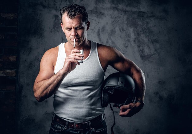 Un homme en t-shirt blanc, fumant une cigarette et tenant un casque de moto dans un studio photo.