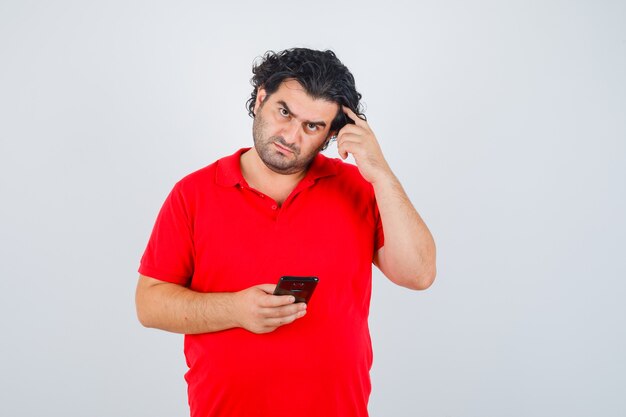 Homme en t-shirt rouge tenant le téléphone, mettant l'index sur la tempe et regardant pensif.