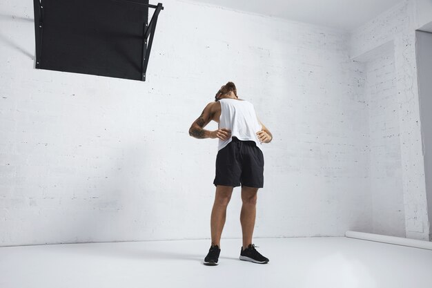 Homme tatoué athlétique en t-shirt réservoir blanc blanc étirant sa poitrine et ses abdominaux après l'entraînement, isolé sur un mur de briques, à côté de la barre de traction noire