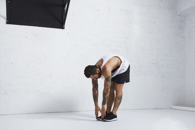 Homme tatoué athlétique en t-shirt réservoir blanc blanc étirant ses jambes en les touchant sur le sol, position du côté gauche