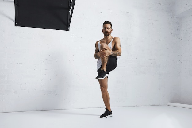 Homme tatoué athlétique en t-shirt réservoir blanc blanc faisant des genoux, étirant ses jambes avec les yeux fermés