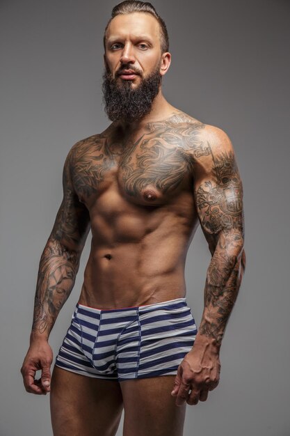Homme tatoué brutal en culotte à rayures posant sur fond gris.