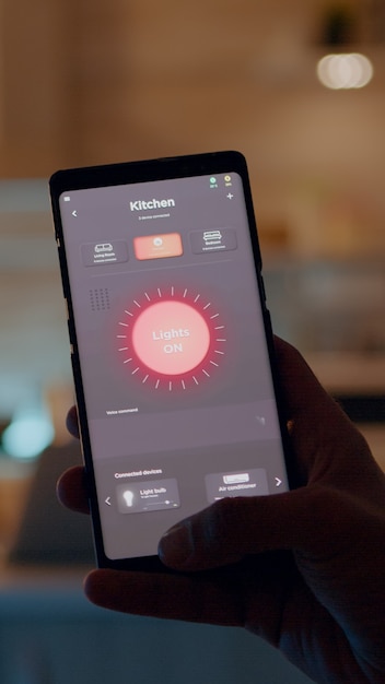 Photo gratuite homme tenant un smartphone avec application de contrôle d'éclairage, allumant les lumières assis dans la cuisine avec système d'éclairage automatisé. personne utilisant un logiciel de maison intelligente travaillant à distance sur un ordinateur portable