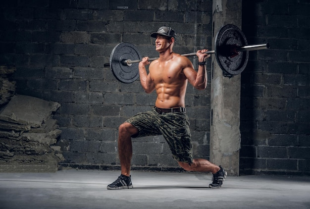 Un homme torse nu attrayant en casquette fait une fente avec une barre au studio photo sombre.