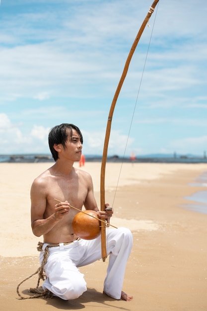 Photo gratuite homme torse nu pratiquant la capoeira sur la plage avec un arc en bois