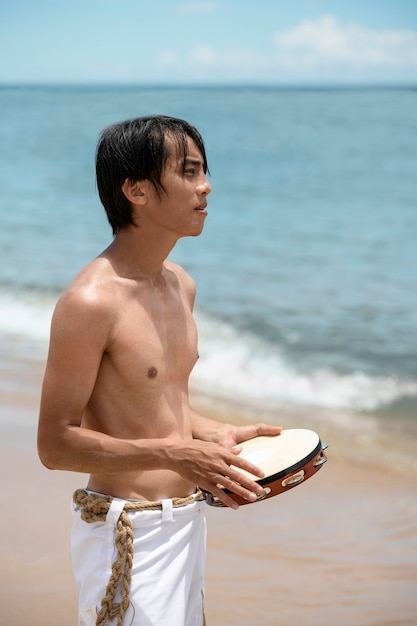 Photo gratuite homme torse nu pratiquant la capoeira sur la plage avec tambourin