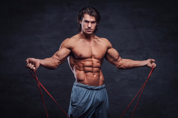 Photo gratuit un homme torse nu séduisant regarde la caméra tout en faisant des exercices avec du caoutchouc. il y a un fond sombre.