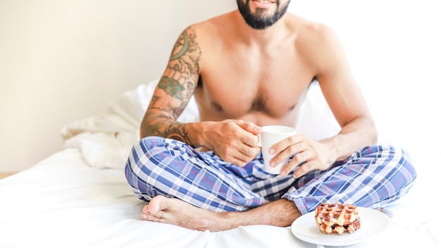 Homme torse nu avec une tasse de café et gaufres assis sur le lit