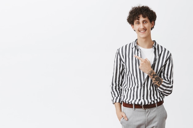 Homme urbain détendu et sympathique avec des tatouages de moustache et des cheveux bouclés pointant vers le coin supérieur gauche souriant avec un regard heureux et ravi dans une chemise cool à rayures