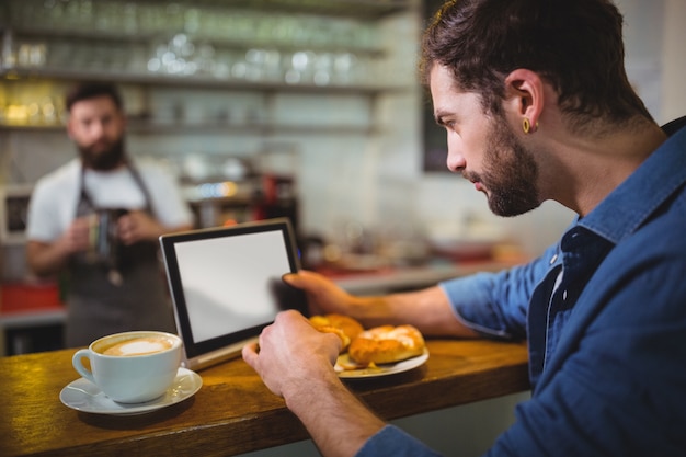 Homme utilisant tablette numérique tout en ayant un croissant dans un café ©