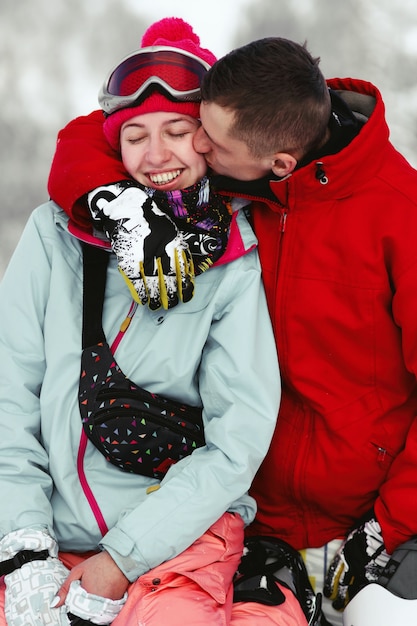 Photo gratuite homme en veste de ski rouge embrasse la joue de la femme alors qu'ils sont assis sur une colline enneigée