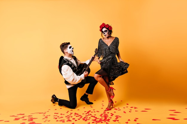 homme zombie jouant à la sérénade. Photo intérieure d'un mec avec du maquillage muerte posant avec une guitare à côté d'une femme en robe noire.