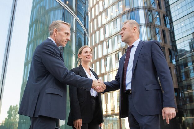 Hommes d'affaires d'âge moyen confiants se saluant et se serrant la main. Jeune femme d'affaires de contenu réussi debout près d'eux et souriant. Construire sur fond. Concept de travail d'équipe et de partenariat