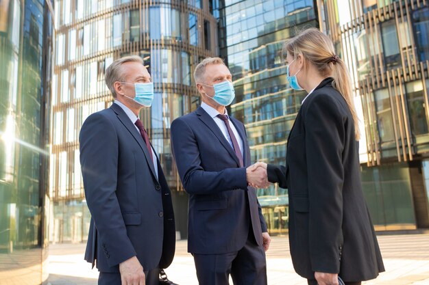 Hommes D'affaires Et Femme Dans Des Masques Faciaux Se Serrant La Main Près Des Immeubles De Bureaux, Se Réunissant Et Parlant En Ville. Vue Latérale, Faible Angle. Concept De Coopération Et De Coronavirus