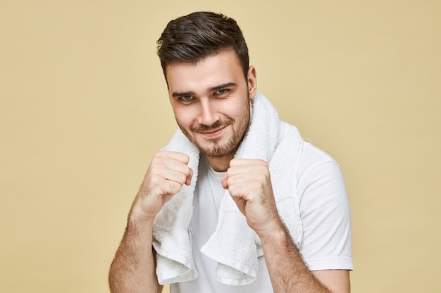 Les hommes et la masculinité. Portrait of young brunette male non rasé avec une serviette autour du cou debout dans la salle de bain tenant les poings serrés devant lui, défiant son propre reflet dans le miroir