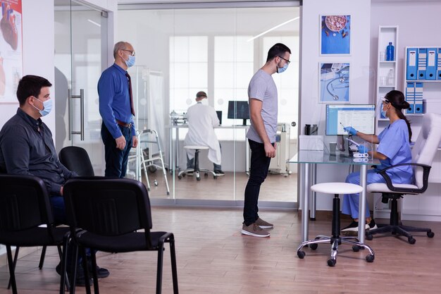 Hommes avec des masques de protection visitant la clinique de l'hôpital, vérifiant le rendez-vous en respectant la distance sociale dans la salle d'attente pendant la pandémie mondiale