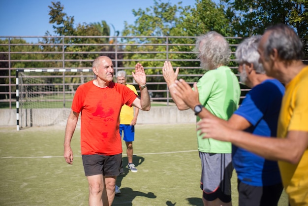 Hommes seniors actifs se remerciant après le match. Hommes aux cheveux gris en vêtements de sport, debout en rang sur le terrain de sport, donnant des high fives. Football, sport, concept de loisirs