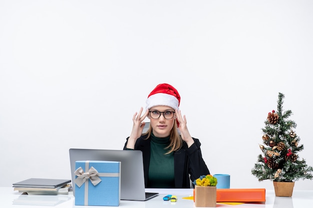 L'humeur du nouvel an avec une femme déterminée avec un chapeau de père Noël assis à une table avec un arbre de Noël et un cadeau sur elle sur fond blanc