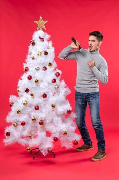 L'humeur du nouvel an avec un gars positif émotionnel chantant la chanson debout près de l'arbre de Noël décoré sur le rouge