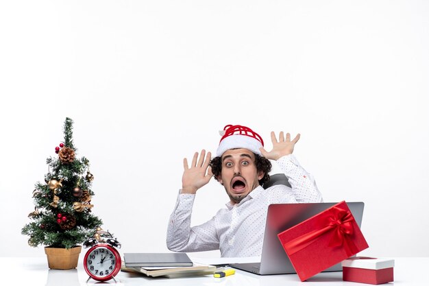 L'humeur du nouvel an avec un jeune homme d'affaires nerveux choqué avec un chapeau de père Noël assis dans le bureau et travaillant seul sur un projet sur fond blanc