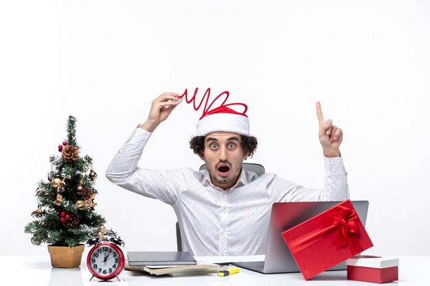 L'humeur du nouvel an avec surpris jeune homme d'affaires touchant son drôle de chapeau de père Noël pointant ci-dessus dans le bureau sur fond blanc