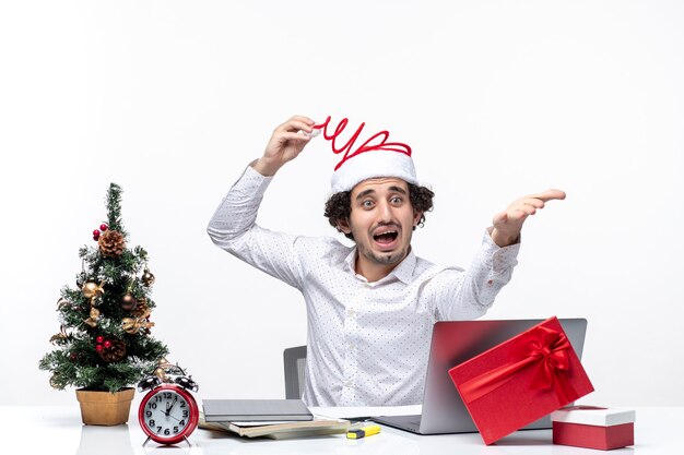 L'humeur du nouvel an avec surpris souriant jeune homme d'affaires touchant son drôle de chapeau de père Noël interrogeant quelque chose dans le bureau sur fond blanc