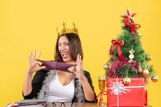 Humeur de Noël avec belle dame en costume avec couronne tenant son masque médical au bureau sur jaune