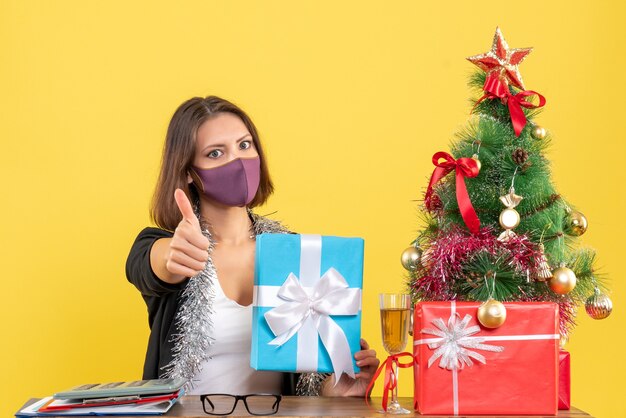 Humeur de Noël avec belle dame en costume avec masque médical et tenant un cadeau au bureau en faisant un geste ok sur jaune