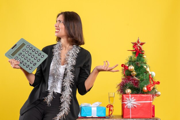 Humeur de Noël avec une belle dame nerveuse debout dans le bureau et tenant la calculatrice au bureau sur jaune