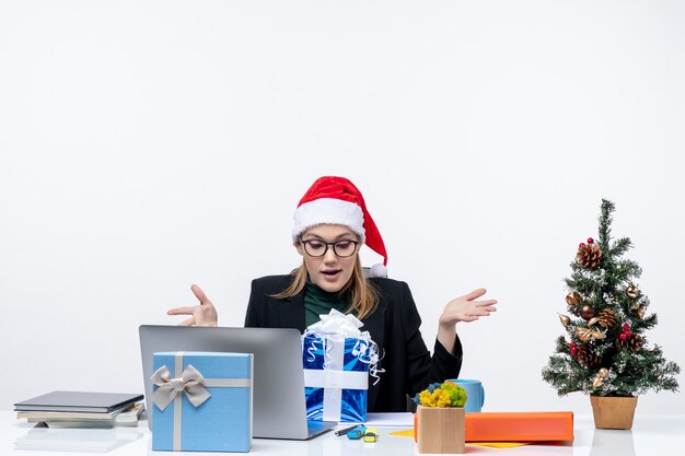 L'humeur de Noël avec excité jeune femme avec chapeau de père Noël et portant des lunettes assis à une table en regardant cadeau étonnamment sur fond blanc