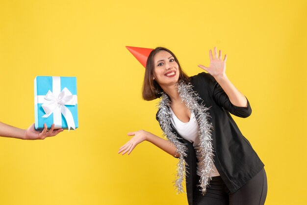 L'humeur de Noël avec une femme d'affaires positive en costume avec un chapeau de Noël et une main tenant un cadeau sur le jaune