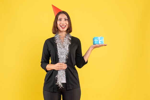 Humeur de Noël avec une femme d'affaires positive en costume avec un chapeau de Noël et tenant un cadeau sur jaune