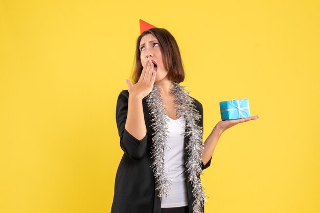 L'humeur de Noël avec une femme d'affaires surprise choquée en costume avec chapeau de Noël et tenant un cadeau sur jaune