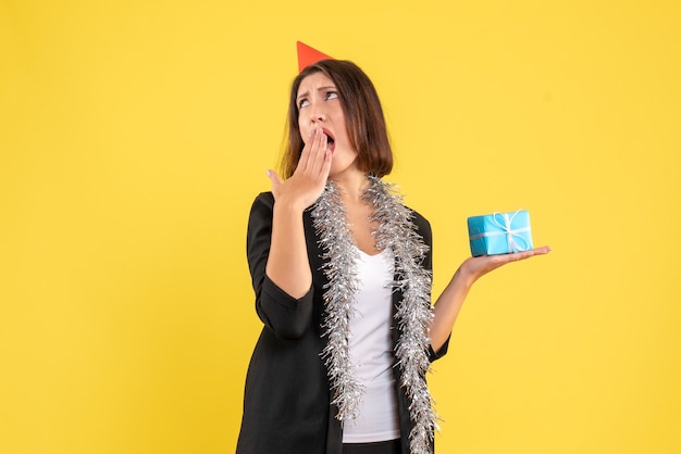 L'humeur de Noël avec une femme d'affaires surprise choquée en costume avec chapeau de Noël et tenant un cadeau sur jaune