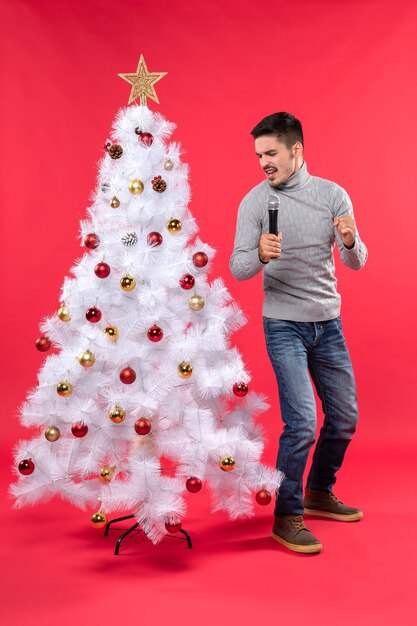 L'humeur de Noël avec un gars confiant habillé en jeans debout près de l'arbre de Noël décoré et tenant le chant du microphone