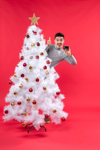 L'humeur de Noël avec un gars émotionnel debout derrière l'arbre de Noël décoré et faisant un geste correct
