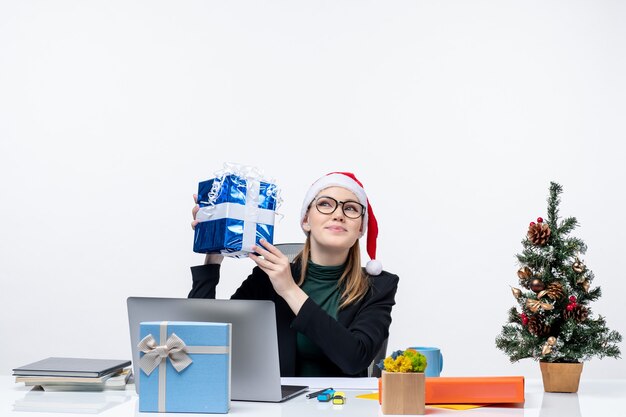 Humeur de Noël avec happy young woman with santa claus hat et portant des lunettes assis à une table tenant son cadeau étonnamment sur fond blanc