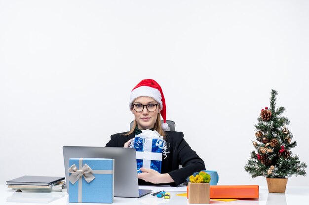 L'humeur de Noël avec jeune femme avec chapeau de père Noël et portant des lunettes assis à une table tenant son cadeau fièrement sur fond blanc