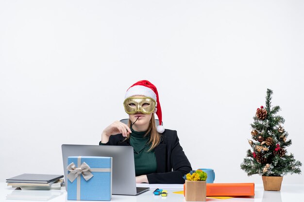 Humeur de Noël avec jeune femme confiante avec chapeau de père Noël tenant des lunettes et portant un masque assis à une table sur fond blanc