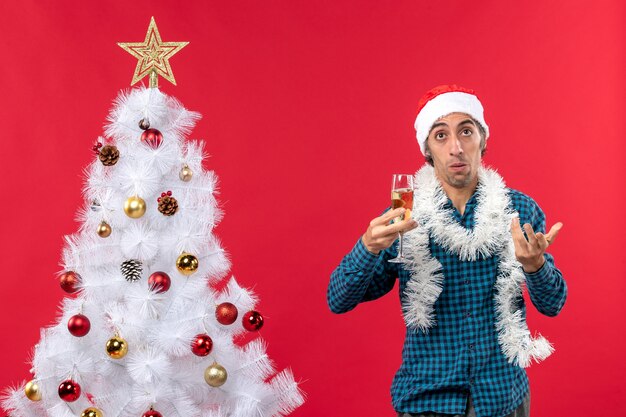 L'humeur de Noël avec jeune homme concerné avec chapeau de père Noël dans une chemise dépouillé bleu soulevant un verre de vin près de l'arbre de Noël