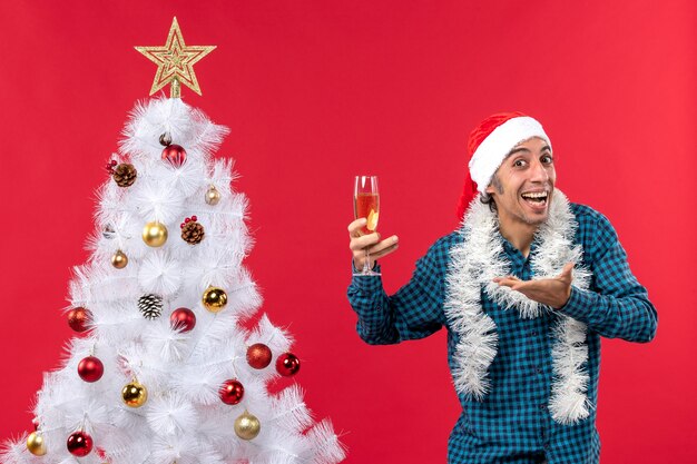 L'humeur de Noël avec jeune homme émotionnel avec chapeau de père Noël dans une chemise bleue dépouillée tenant un verre de vin et demandant quelque chose près de l'arbre de Noël