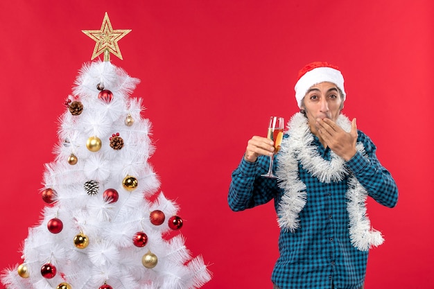 L'humeur de Noël avec jeune homme émotionnel avec chapeau de père Noël dans une chemise bleue dépouillée tenant un verre de vin en mettant les doigts