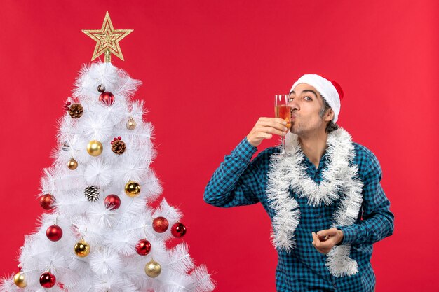 L'humeur de Noël avec jeune homme émotionnel avec chapeau de père Noël dans une chemise dépouillé bleu tenant et embrassant le verre avec du vin près de l'arbre de Noël