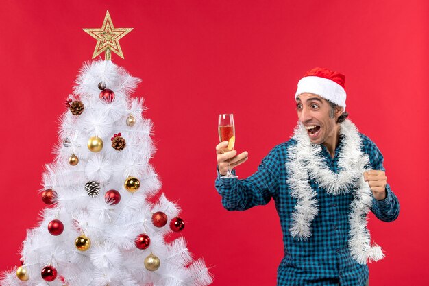 L'humeur de Noël avec un jeune homme émotionnel fou avec un chapeau de père Noël dans une chemise rayée bleue tenant un verre de vin près de l'arbre de Noël